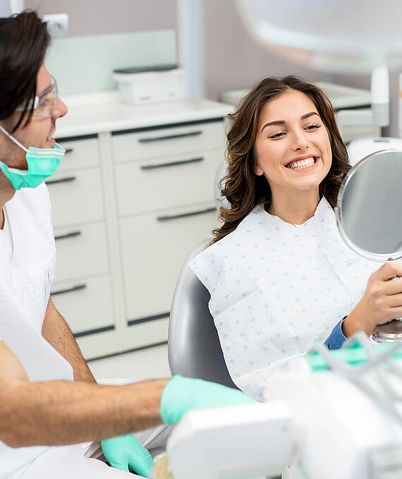 A young woman is smiling as she examines her teeth in a mirror. She is sitting in a dentist’s chair. A dentist is sitting next to her.